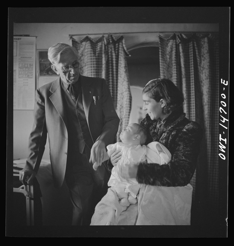 Woman sitting in a room with a baby in her lap. Older man is standing holding the baby's hand and looking at the baby. Baby is looking at the man. Woman is looking at the baby.