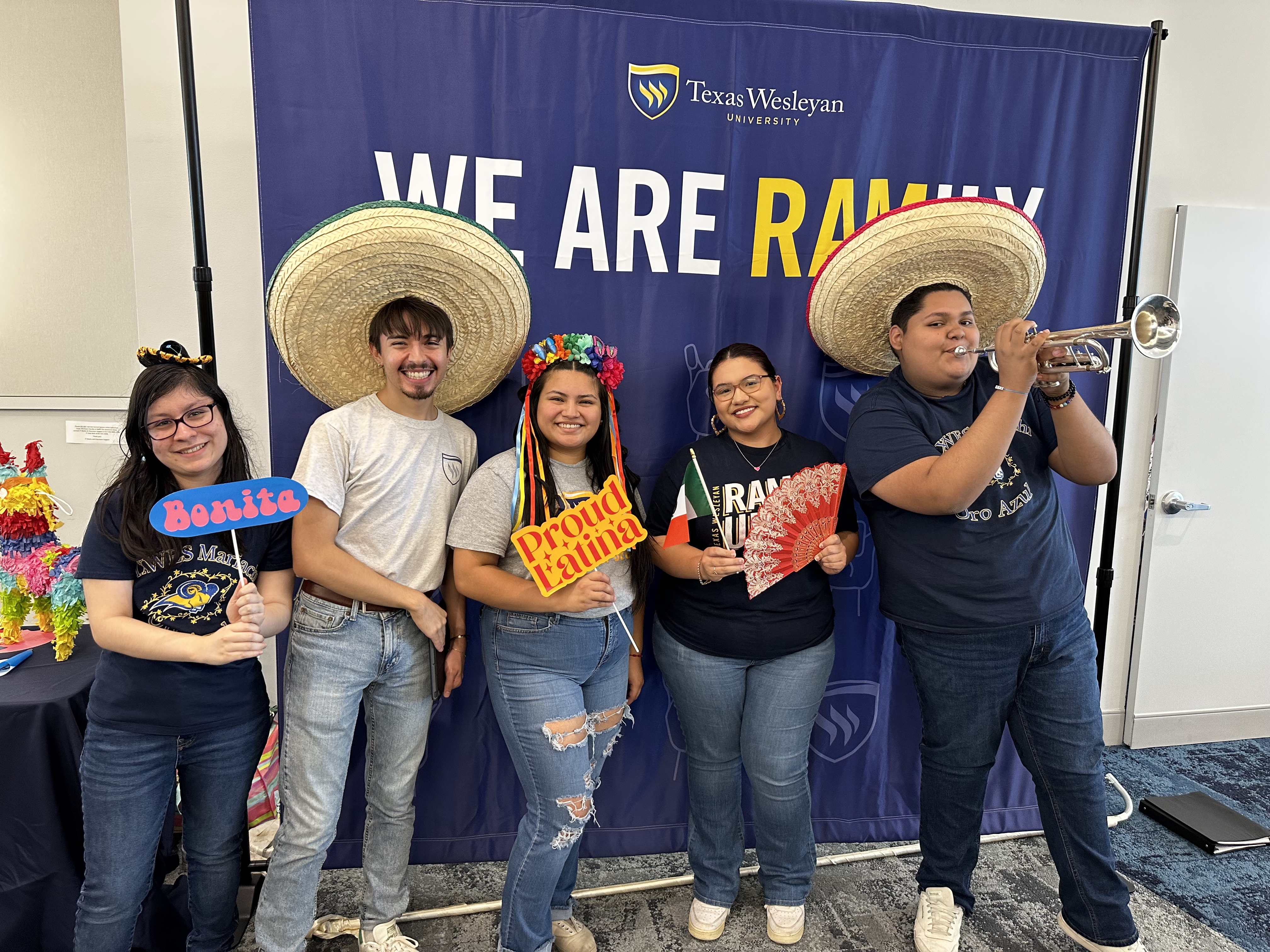 Students pose with trumpet, sombreos, and signs in front of a banner that says We Are Ramily