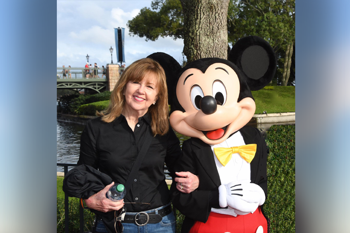 A photo of Linda Metcalf wearing a black blouse next to Mickey Mouse in a tuxedo with a yellow bow tie.