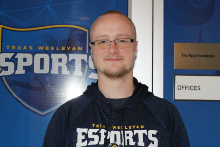 Young male college student stands in front of a door with the Texas Wesleyan esports logo on it